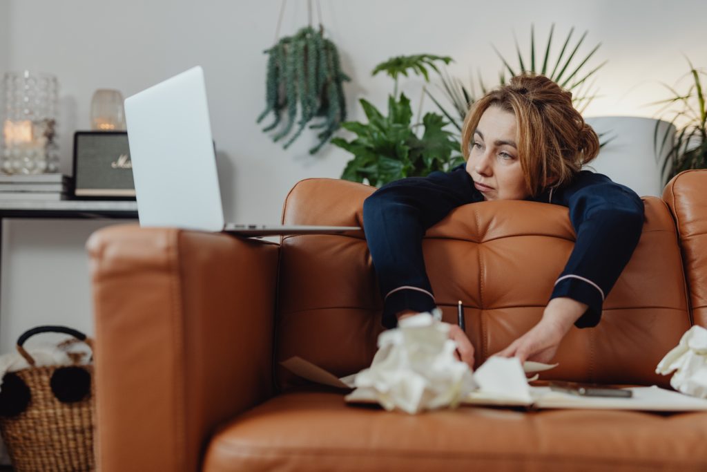 Woman lying on a brown leather sofa, looking at her laptop with a thoughtful expression, surrounded by crumpled papers and a notebook as she prepares to restore from a backup.