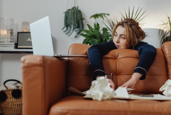 Woman lying on a brown leather sofa, looking at her laptop with a thoughtful expression, surrounded by crumpled papers and a notebook as she prepares to restore from a backup.