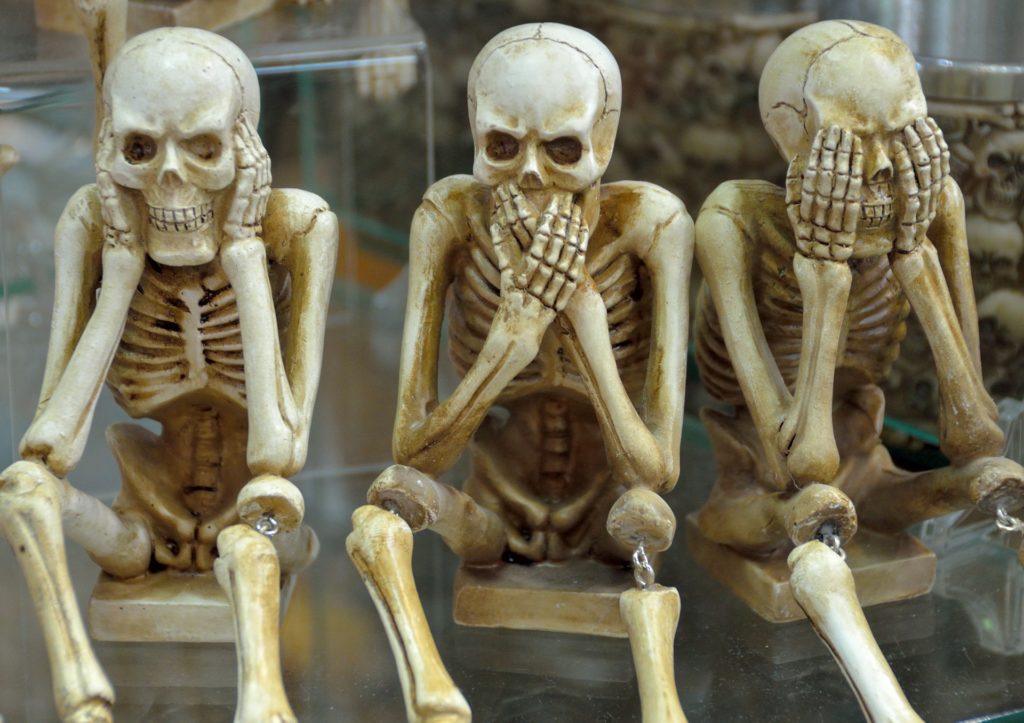 Three backup skeleton figurines sitting, each covering its face with hands in a "see no evil, hear no evil, speak no evil" pose.