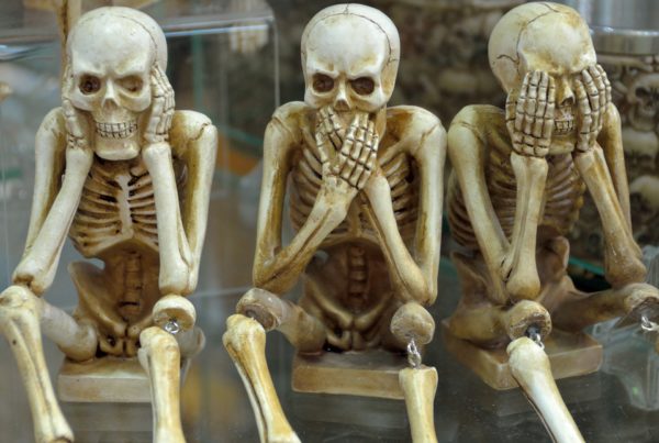 Three backup skeleton figurines sitting, each covering its face with hands in a "see no evil, hear no evil, speak no evil" pose.