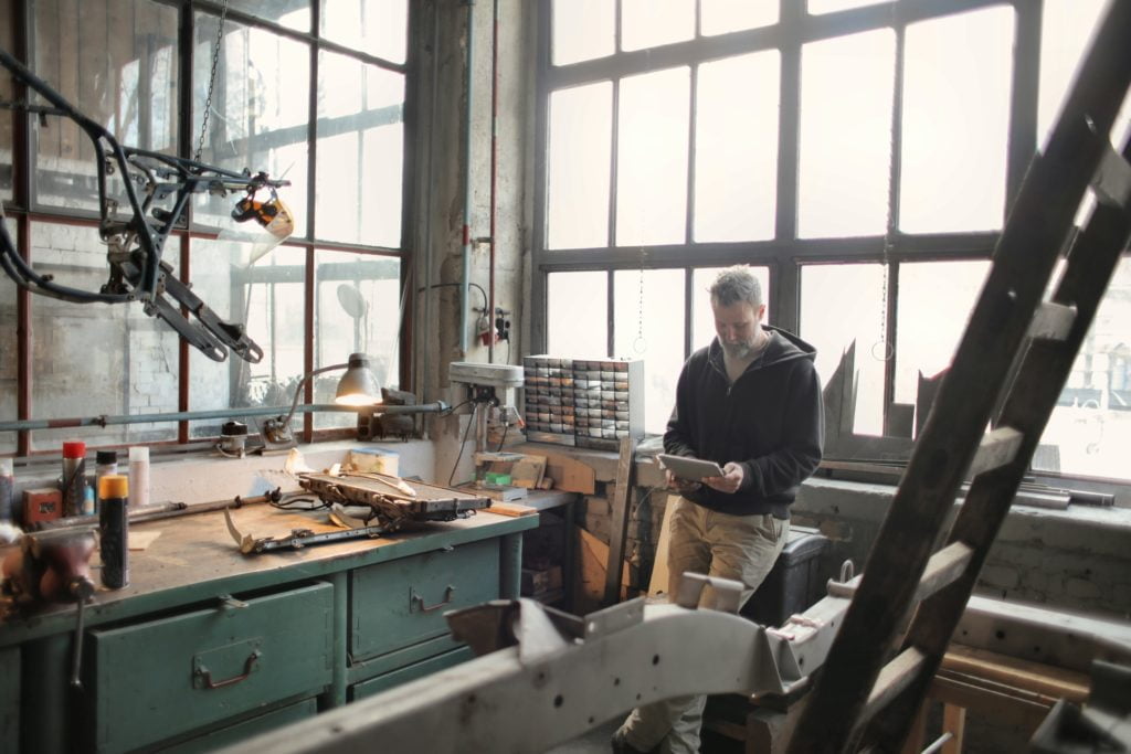 A man reviews notes on a clipboard in a cluttered workshop with tools and an Azure Information Protection-secured robotic arm near large windows.