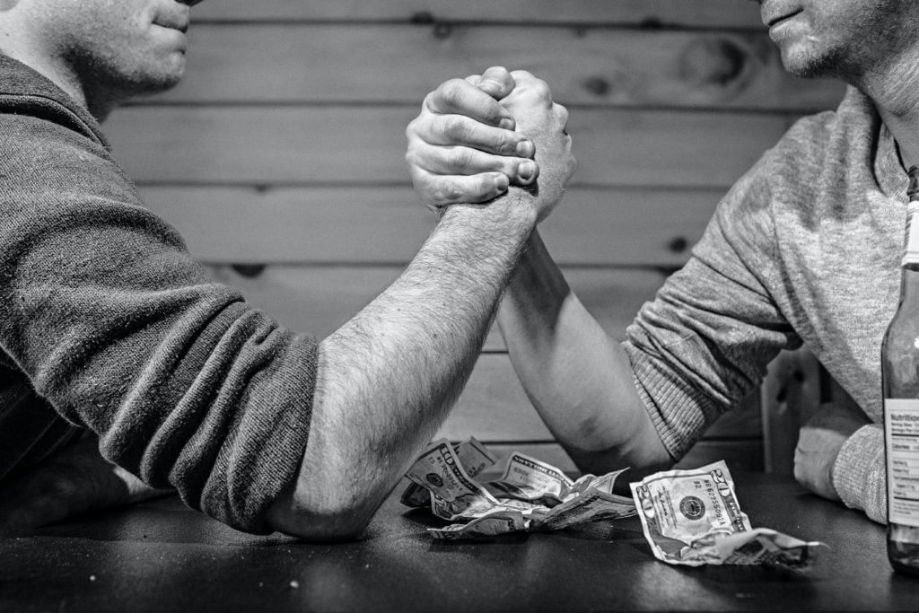 Two men engaging in an arm wrestling match over a table scattered with dollar bills.