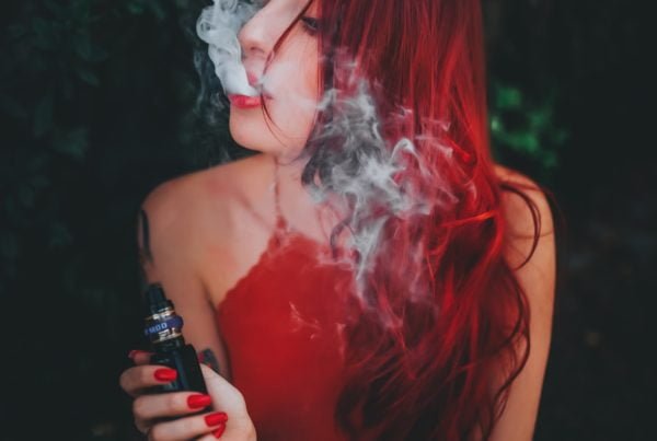 A woman with red hair exhaling smoke while holding a vape device with a built-in vape detector, sitting against a leafy background.