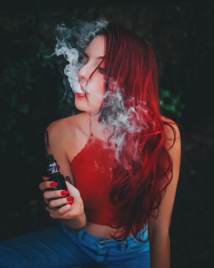 A woman with red hair exhaling smoke while holding a vape device with a built-in vape detector, sitting against a leafy background.