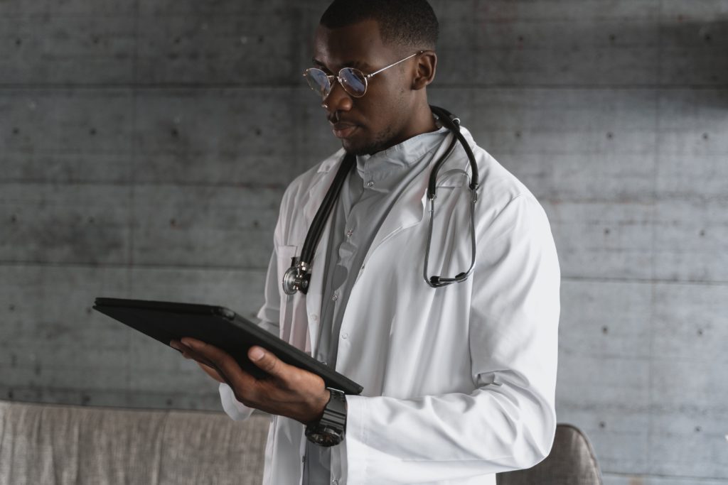 Black male doctor in a white coat and stethoscope reading a digital tablet with Azure Information Protection in a modern office setting.