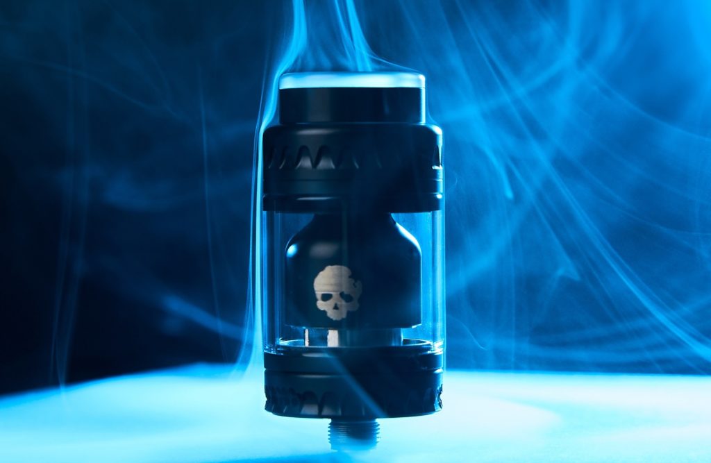 A black, futuristic-looking mechanical mod vape with a visible skull design inside, surrounded by swirls of blue vapor against a dark background.