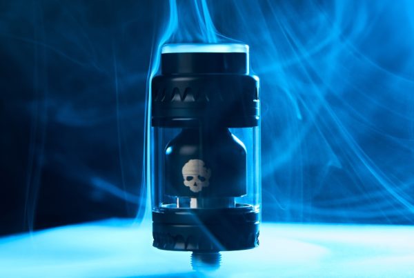 A black, futuristic-looking mechanical mod vape with a visible skull design inside, surrounded by swirls of blue vapor against a dark background.