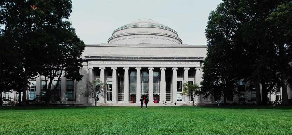Frontal view of the massachusetts institute of technology's great dome and entrance, flanked by trees, with people walking in front.