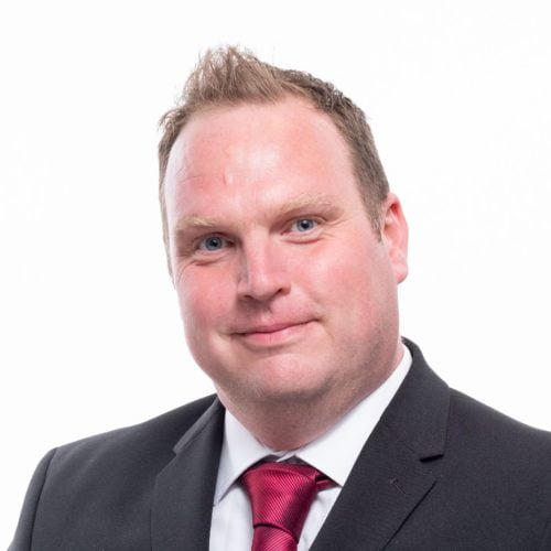 Professional portrait of a man in a suit with a red tie, smiling at the camera against a white background for our "Meet the Team" section.