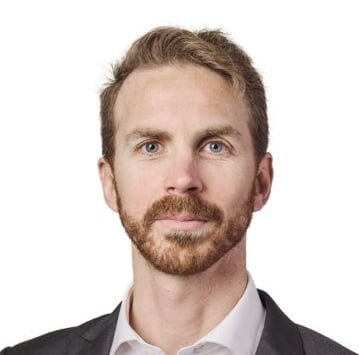 Meet the team: A professional headshot of a man with a light beard and short brown hair, wearing a dark suit and white shirt.