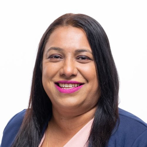 Meet the team: Portrait of a smiling woman with long black hair and bright pink lipstick, wearing a blue blazer against a white background.