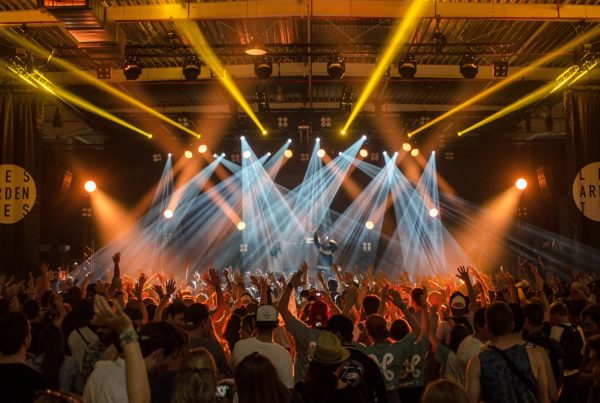 Crowd of people enjoying a concert with hands up, vibrant stage lights illuminating the venue from above, all compliant with Martyn's Law security measures.